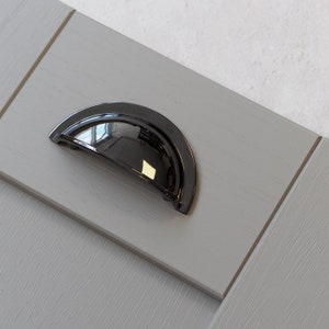 May include: A black cup-shaped cabinet pull with a shiny finish. The pull is mounted on a light gray wooden surface.