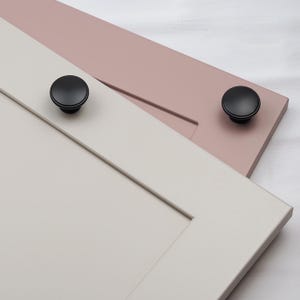 Puede incluir: Dos puertas de armario con pomos redondos negros. Una puerta está pintada de color rosa claro y la otra de color gris claro.