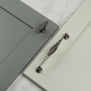May include: A dark gray cabinet door with a small, spiraled, metallic knob and a light sage green cabinet door with a matching, elongated, spiraled, metallic handle. Both pieces of hardware feature a dark, weathered, antique-style finish.