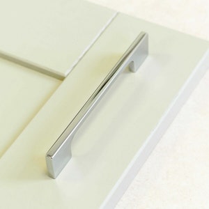 May include: A chrome cabinet pull with a rectangular shape and a slight curve. The pull is mounted on a white cabinet door.
