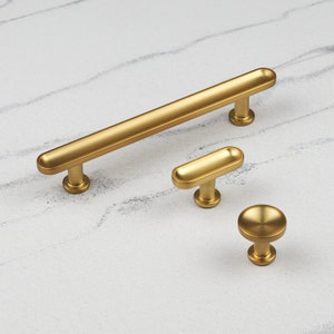 May include: Three gold-toned cabinet hardware pulls in different sizes and styles. The largest pull is a long, rectangular bar with rounded edges. The medium pull is a smaller, rectangular bar with rounded edges. The smallest pull is a round knob with a flat top.