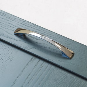 Peut inclure: Une poignée de meuble chromée avec un design incurvé. La poignée est montée sur une porte de meuble en bois bleu.