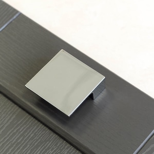 Peut inclure: Une poignée de tiroir carrée en chrome avec un bord légèrement surélevé. La poignée est montée sur une porte de placard gris foncé.