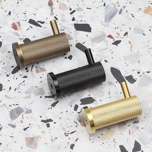 Może przedstawiać: Trzy metalowe haczyki ścienne w kolorze mosiądzu, czarnym i złotym. Każdy haczyk ma cylindryczny korpus z rombowym wzorem i małe, kątowe ramię. Haczyki są prezentowane na nakrapianej, jasnej powierzchni.