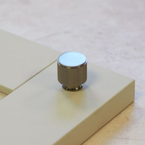 Puede incluir: Un pomo de metal plateado con una superficie texturizada. El pomo es redondo y tiene una parte superior plana.