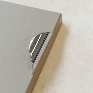 以下が含まれることがあります： 丸みを帯びたエッジを持つクロムメッキの湾曲したキャビネットプル。プルはライトグレーの表面に取り付けられています。