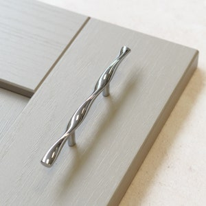 May include: A silver twisted cabinet pull with a smooth, polished finish. The pull is mounted on a light gray cabinet door.