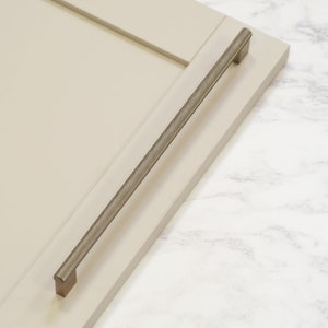 May include: A brushed nickel cabinet pull with a textured finish. The pull is mounted on a light beige cabinet door.