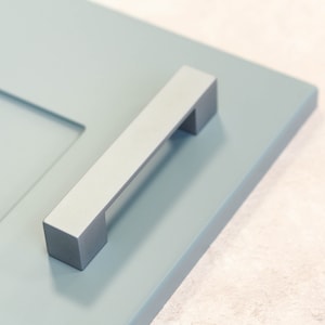 Puede incluir: Una manija de puerta de armario de plata con forma rectangular y un borde ligeramente redondeado. La manija está montada en una puerta de armario azul claro.