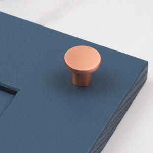 Op de afbeelding: Een ronde koperen kastknop met een geborstelde afwerking. De knop is gemonteerd op een blauwe kastdeur.