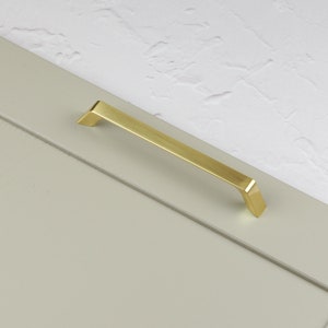 May include: A brushed gold cabinet pull with a rectangular shape. The pull has a slightly angled design at each end.