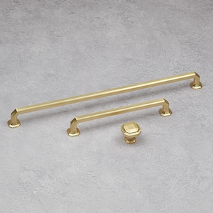May include: A set of three brass-toned cabinet hardware pieces. The set includes a long bar pull, a shorter pull, and a square knob. The hardware has a geometric design and is displayed on a light gray background.