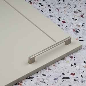 May include: A brushed nickel cabinet pull with a rectangular shape. The pull is mounted on a white cabinet door.