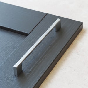 Puede incluir: Un tirador de armario plateado de forma rectangular con un acabado liso y cepillado. El tirador está montado en una puerta de armario gris oscuro.