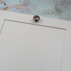 Peut inclure: Gros plan sur une porte d'armoire blanche avec un bouton de couleur bronze. Le bouton est rond et l&eacute;g&egrave;rement bomb&eacute;. L'arri&egrave;re-plan est une surface textur&eacute;e et multicolore.