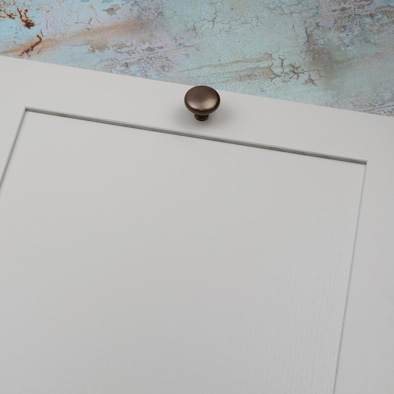 Peut inclure: Gros plan sur une porte d'armoire blanche avec un bouton de couleur bronze. Le bouton est rond et l&eacute;g&egrave;rement bomb&eacute;. L'arri&egrave;re-plan est une surface textur&eacute;e et multicolore.