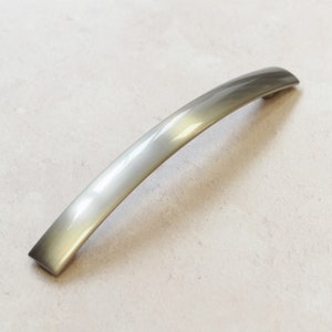 Może przedstawiać: Srebrny, zakrzywiony uchwyt do szafki o gładkim, polerowanym wykończeniu.