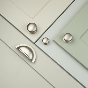 Può includere: Un set di ferramenta per mobili in nichel spazzolato che include due pomelli rotondi e una maniglia a conchiglia semicircolare. La ferramenta è montata su tre ante di armadi separate con finiture color crema, grigio chiaro e verde salvia.