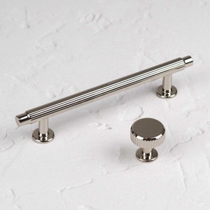 May include: A set of two silver cabinet hardware pieces. The first piece is a 12.7 cm long pull with a ribbed design. The second piece is a round knob with a ribbed design.