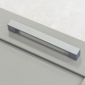 Puede incluir: Un tirador de armario plateado con forma rectangular y bordes ligeramente redondeados. El tirador está montado en una puerta de armario gris.