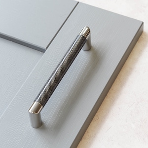 Op de afbeelding: Een zilveren kastdeurgreep met een zwart, getextureerd ontwerp. De greep is gemonteerd op een grijze kastdeur.