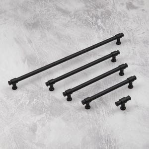 以下が含まれることがあります： さまざまな長さの、マットブラックのキャビネットプル5個セット。各プルは、丸みを帯びたエンドキャップと装飾的なベースを備えた円筒形のバーを備えています。ハードウェアは、明るい灰色のテクスチャード加工された表面に斜めに配置されています。