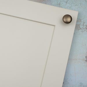 Peut inclure: Gros plan sur une section d'une porte d'armoire blanche avec un bouton de couleur bronze. La porte pr&eacute;sente un design &agrave; panneaux encastr&eacute;s et une esth&eacute;tique simple et moderne. Le bouton est rond et a une finition bross&eacute;e.