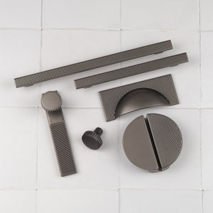 May include: A set of seven modern cabinet hardware pulls in a dark gray finish. The pulls feature a variety of shapes and sizes, including a round knob, a rectangular pull, and a curved pull.