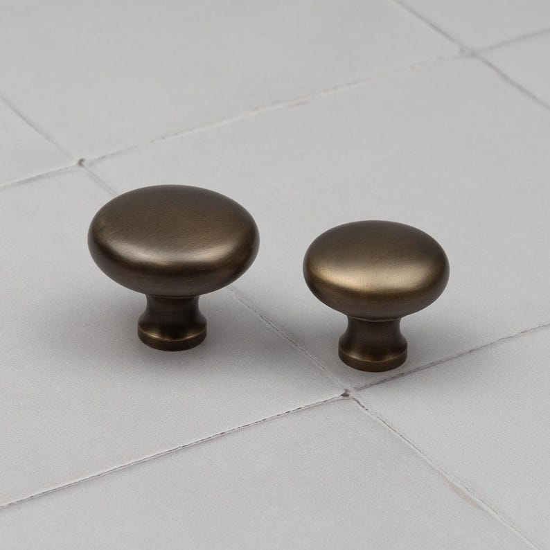Peut inclure: Deux boutons de meuble en bronze avec un dessus arrondi et une base cylindrique courte. Les boutons sont pr&eacute;sent&eacute;s sur une surface gris clair, mettant en valeur leur finition m&eacute;tallique et leur design simple.