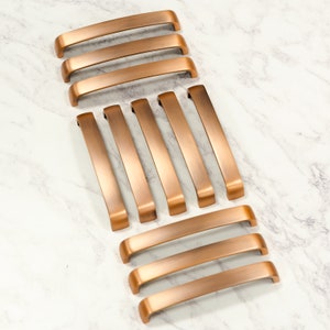 May include: A set of 10 brushed copper cabinet pulls. The pulls are arranged in two rows, with five pulls in each row. The pulls are all the same size and shape, and they have a slightly rounded edge.