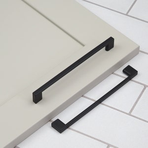 Puede incluir: Dos tiradores de armario negros con forma rectangular. El tirador más grande mide 25,4 cm de largo y el más pequeño 12,7 cm.