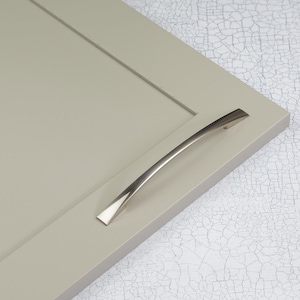 May include: A brushed nickel cabinet pull with a curved design. The pull is mounted on a light grey cabinet door.