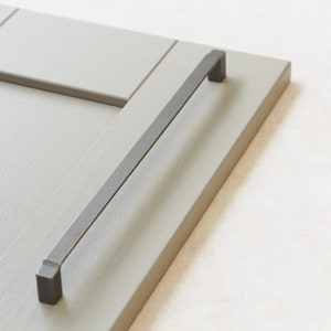 Op de afbeelding: Een close-up van een grijze kastdeur met een zwart metalen handvat. Het handvat is rechthoekig en heeft een licht gebogen vorm.