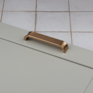 May include: A brushed gold cabinet handle with a curved design. The handle is mounted on a light grey surface.