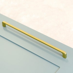 May include: A gold cabinet pull with a rectangular shape. The pull is mounted on a light blue cabinet door.