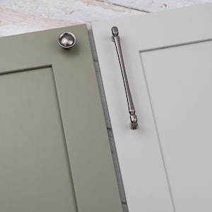 Könnte beinhalten: Nahaufnahme von zwei Schranktüren, eine salbeigrün und eine weiß. Die grüne Tür hat einen runden, gebürsteten Nickelknopf. Die weiße Tür hat einen langen, gebürsteten Nickelgriff. Beide Türen sind vor einem hellen Holzmaserungshintergrund.