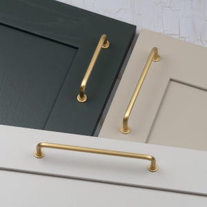 May include: Gold-toned cabinet pulls in various sizes are displayed against a backdrop of painted cabinet doors in shades of gray, green, and beige. The pulls have a sleek, modern design, perfect for kitchen or bathroom renovations.