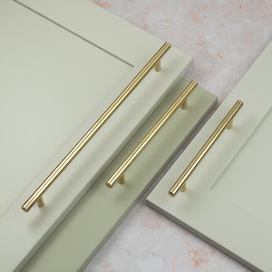 May include: Three cylindrical gold-toned metal cabinet pulls with a smooth, polished finish, each mounted horizontally onto light sage green cabinet doors featuring a recessed center panel design and clean, straight edges.