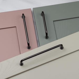 May include: Close-up of cabinet hardware. The image shows three cabinet doors in pastel colours: pink, green, and off-white. Each door has a black, curved handle. The handles are mounted with circular bases. The hardware is a modern design.