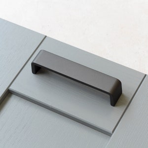 May include: A black metal cabinet pull with a rectangular shape and rounded edges. The pull is mounted on a light gray surface.