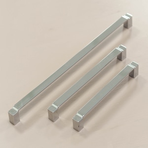 May include: Three silver cabinet pulls of different lengths. The pulls are rectangular with rounded edges and a slightly raised lip on the top and bottom.