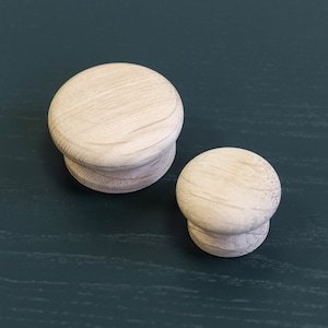 Puede incluir: Dos pomos de madera, uno más grande que el otro, se muestran sobre una superficie verde oscuro. Ambos pomos son redondos y tienen un acabado de madera lisa y clara.