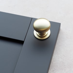 May include: A gold cabinet knob with a round, smooth surface. The knob is mounted on a dark gray cabinet door.