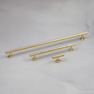 May include: A set of four gold-toned cabinet pulls in varying sizes. The pulls have a ribbed design and a T-bar shape. The hardware is displayed on a light gray surface.
