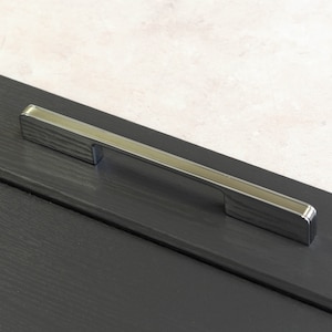 Könnte beinhalten: Ein silberner Schrankgriff mit gebürsteter Oberfläche. Der Griff ist an einer schwarzen Schranktür montiert.