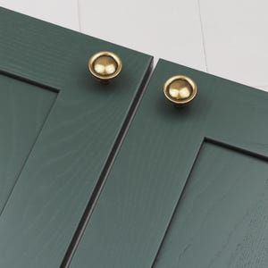 以下が含まれることがあります： 丸みを帯びたドーム型の金色のキャビネットノブが2つ。ノブは濃い緑色の木製のキャビネットドアに取り付けられています。