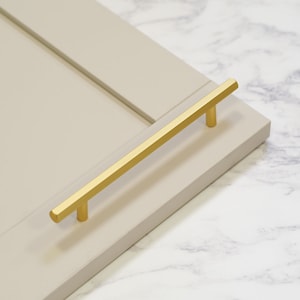 May include: A gold-colored cabinet pull handle with a rectangular bar and two cylindrical posts. The handle is mounted on a light gray cabinet door. The background is a white and gray marble pattern.