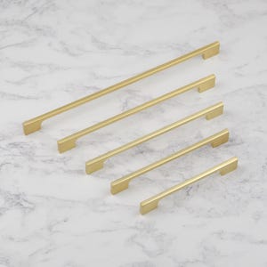 May include: A collection of five gold-colored cabinet pulls in varying lengths. The pulls have a sleek, modern design with a rectangular bar and a flat, rectangular handle. The hardware is arranged diagonally on a white and gray marble surface.