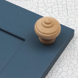 Pode incluir: Um botão de armário de madeira com um design arredondado e canelado. O botão é de cor castanho claro e está montado numa porta de armário pintada de azul.