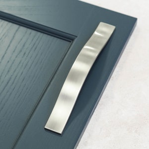 May include: A brushed nickel cabinet door handle with a curved design. The handle is mounted on a dark blue cabinet door.